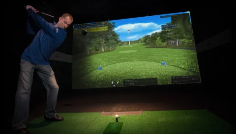 A man takes a swing at X-Golf. On the simulator screen is the Spyglass Hill golf course.