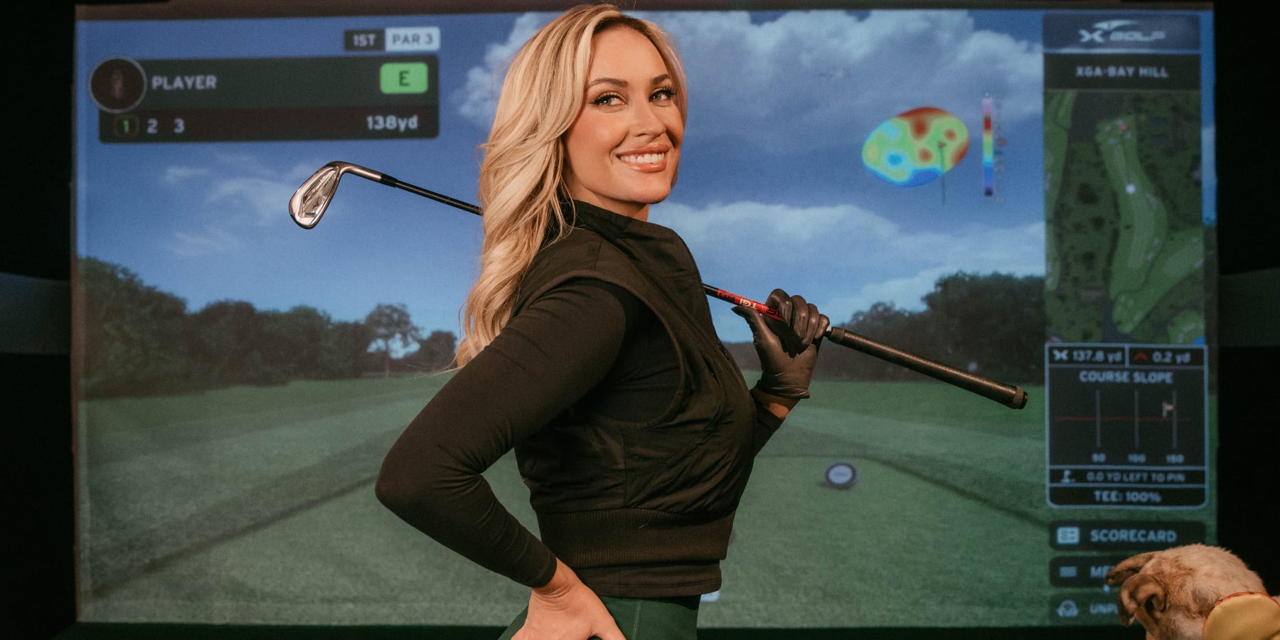 Golfer Paige Spiranac, smiling, stands in front of a golf simulator screen at X-Golf. She stands in profile facing the camera, with her club resting on her shoulder.