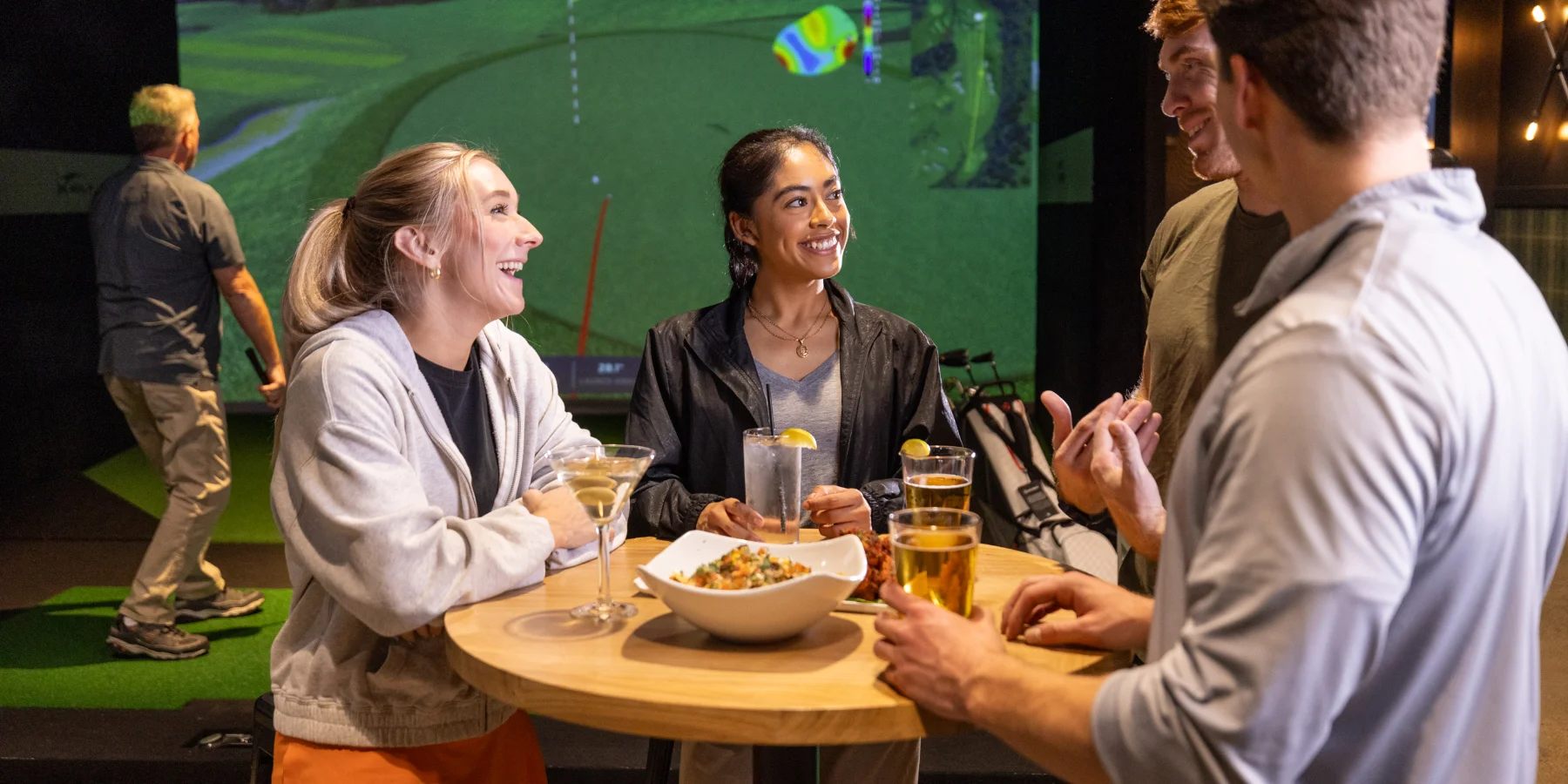 A group of friends smile and laugh while enjoying drinks and appetizers around a table at X-Golf. In the background, a man plays golf at a simulator.